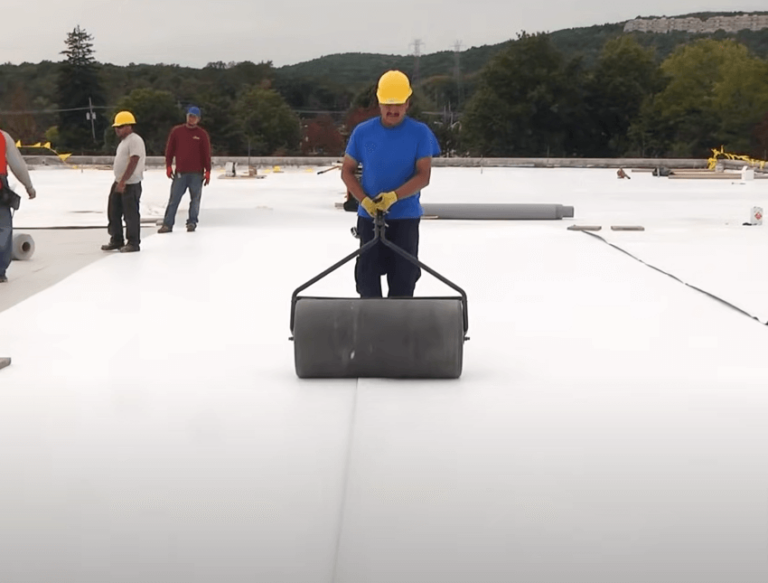 Construction worker rolling a heavy cylinder on a white flat roof, other workers nearby.