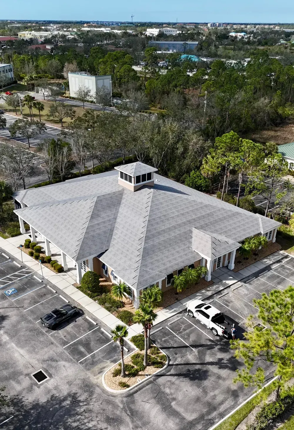 Aerial view of a building with a gray roof and parking lot. Cars are parked around it with trees.