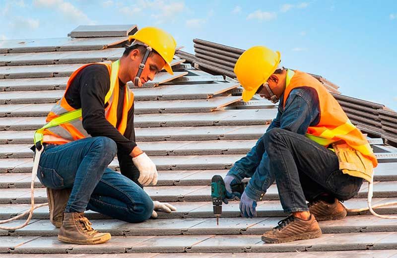 Two construction workers in yellow hard hats installing roof tiles.
