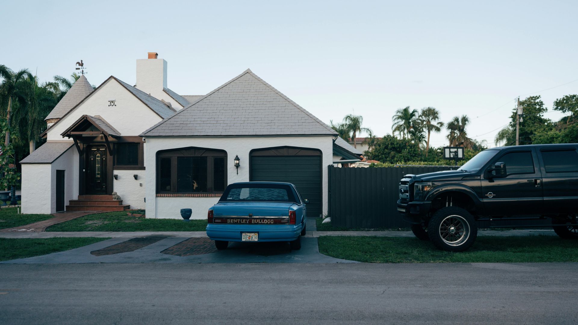 White house with a blue car in the driveway and a large black truck parked on the side.