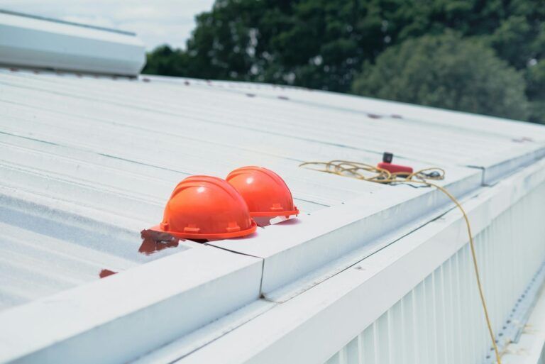 Two orange hard hats and safety equipment sit on a white metal roof's edge.