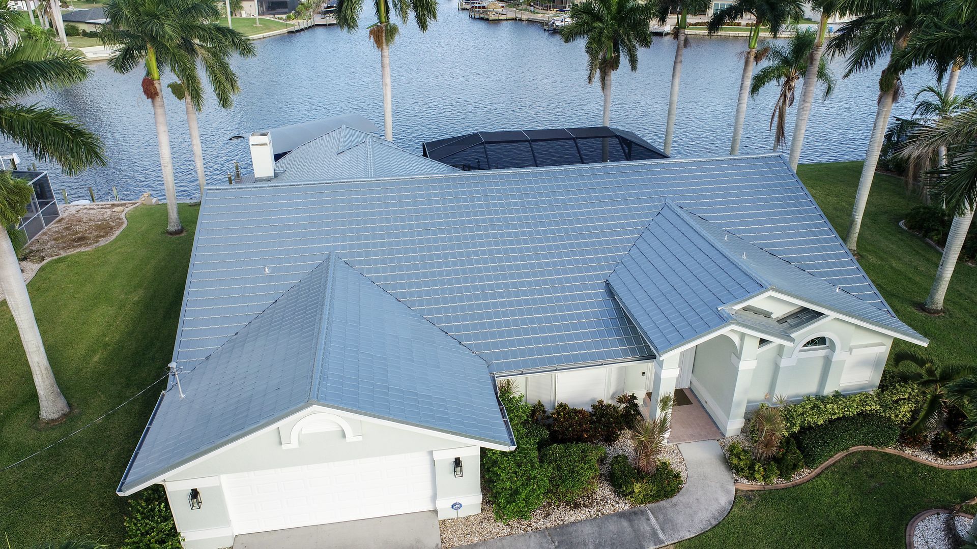 Aerial view of a light blue roofed house with a waterfront view, palm trees, and green grass.