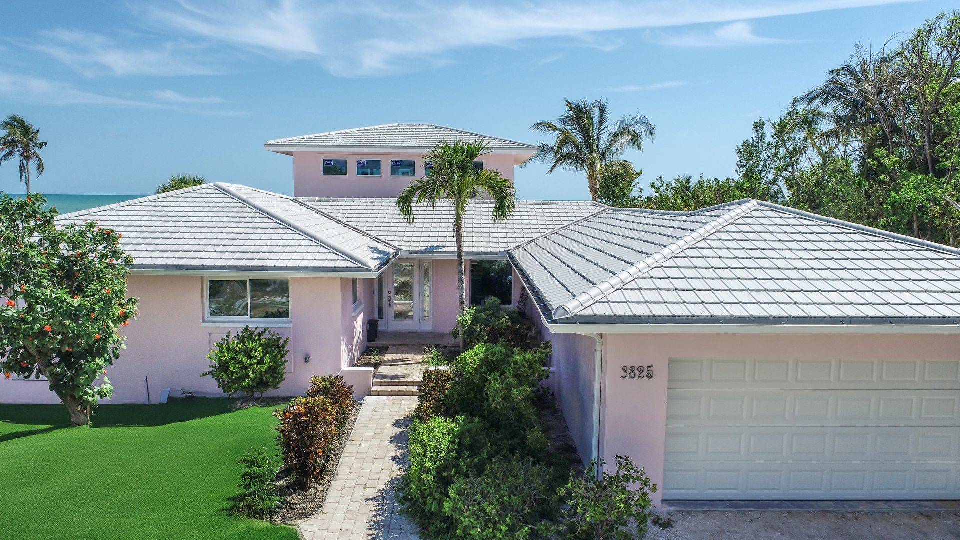 Pink house with white roof, palm trees, and green lawn on a sunny day near the ocean.