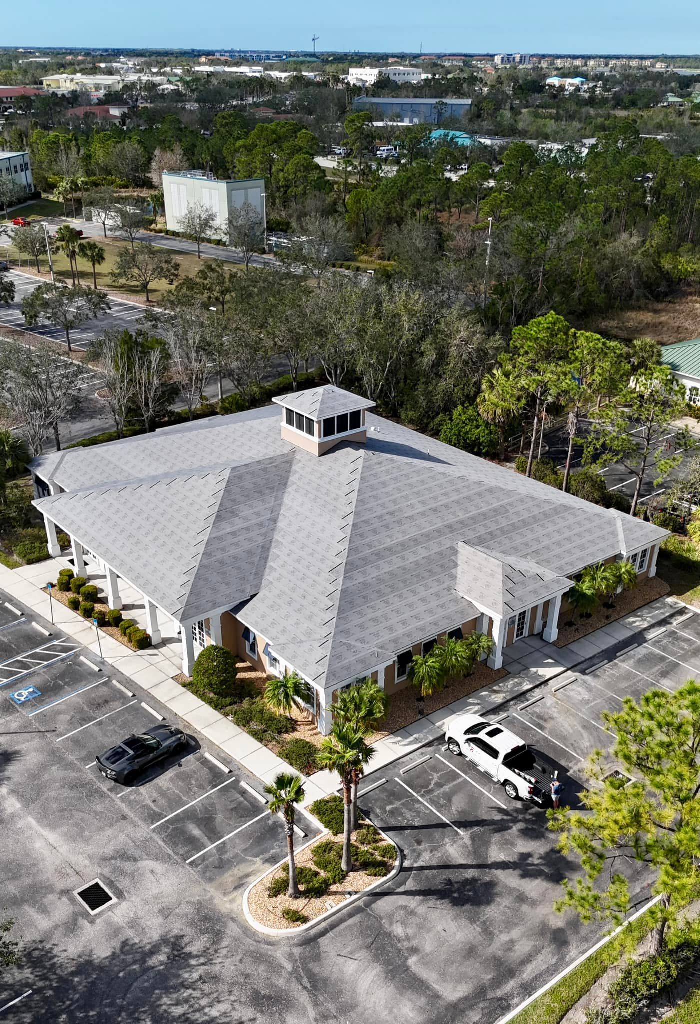 Aerial view of a building with gray roof, white pillars, and a surrounding parking lot with trees in the distance.
