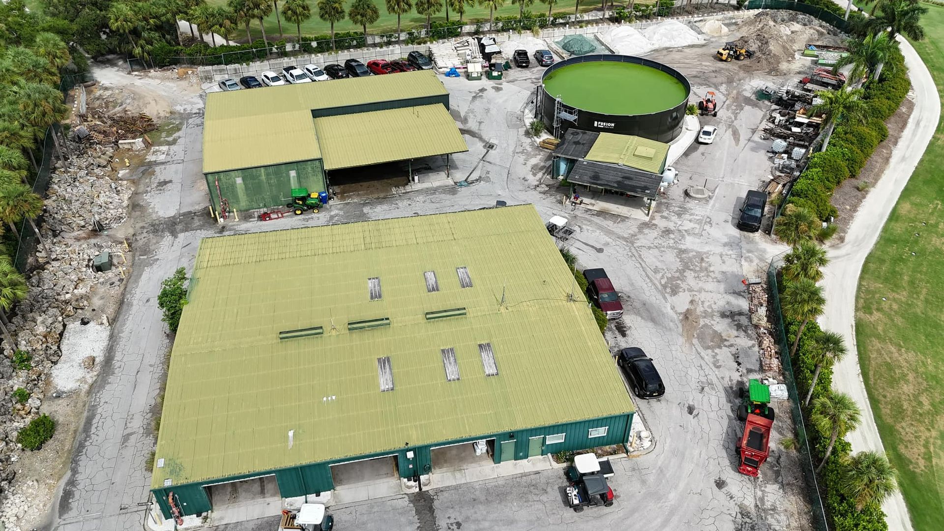 Aerial view of an industrial site with green buildings, a circular green tank, vehicles, and surrounding foliage.
