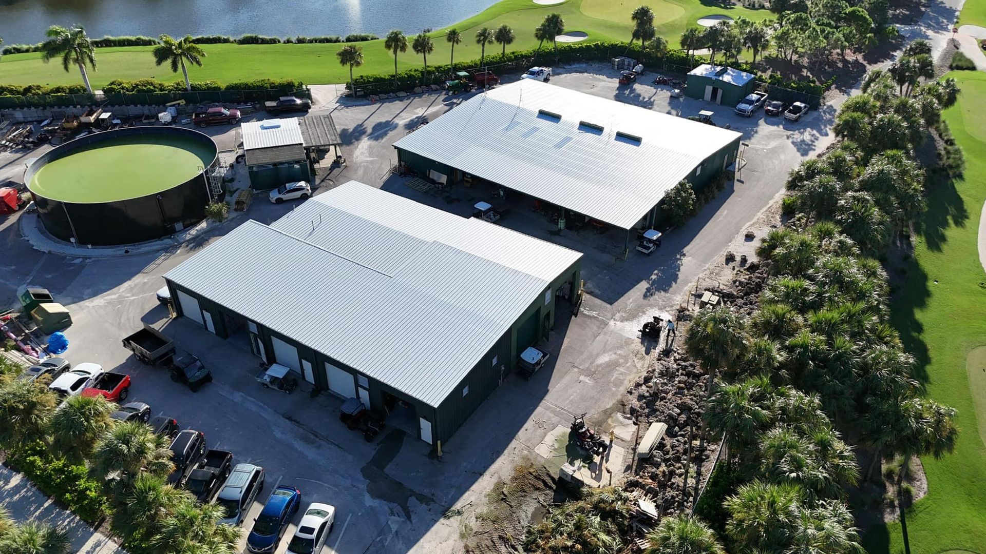Aerial view of two large industrial buildings with vehicles and a water tank, near a golf course.