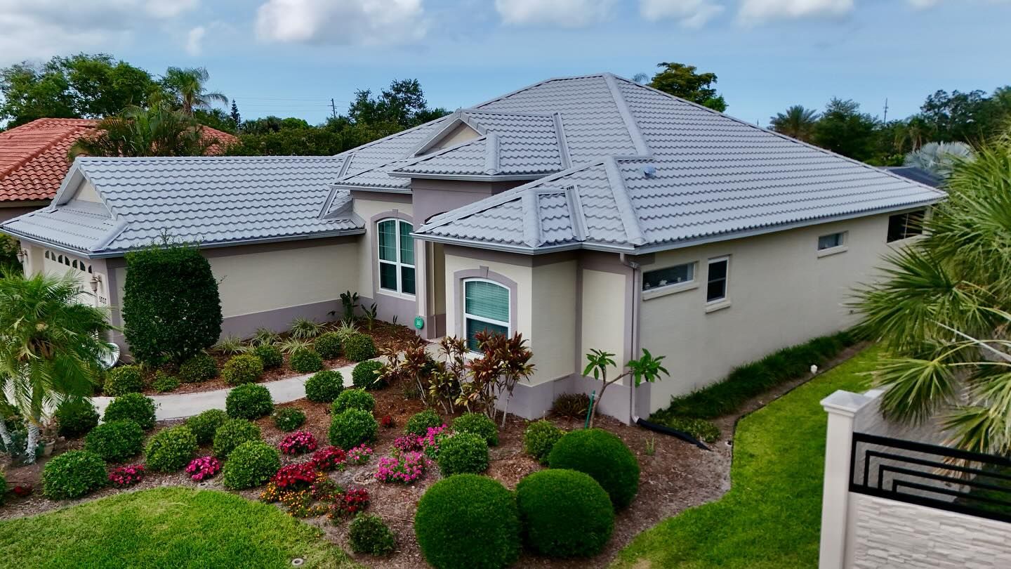 A light-colored house with a gray tiled roof, surrounded by landscaping and trees.