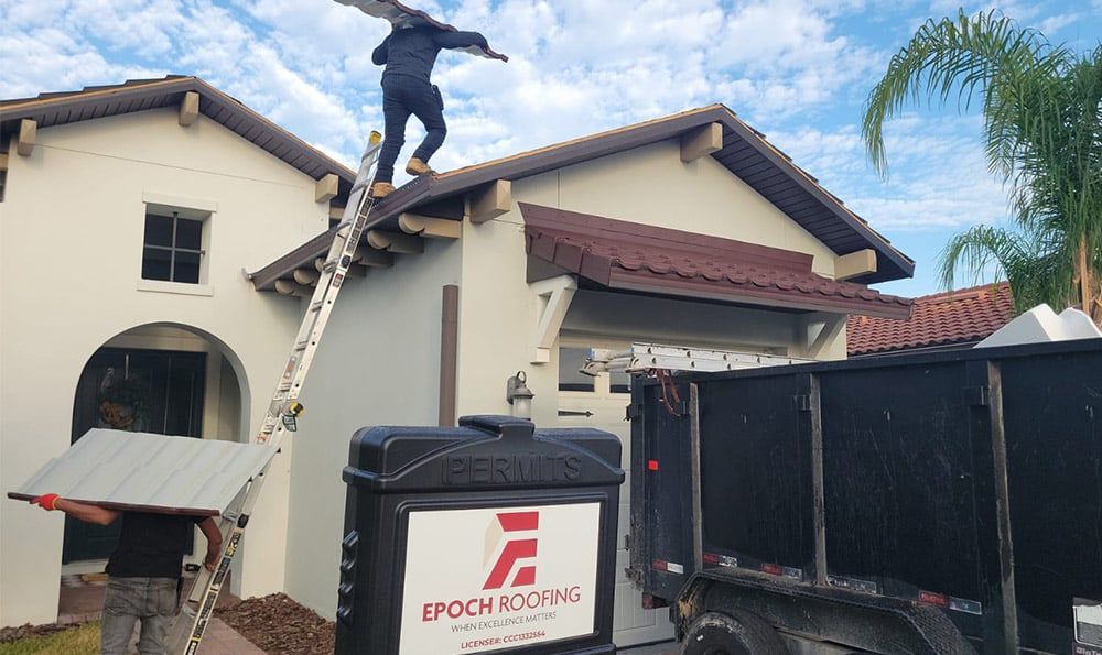 Roofers on a house, one on ladder, one on roof. Roofing company logo on a container, blue sky.