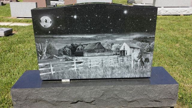 A black and white gravestone with a painting of a farm at night.