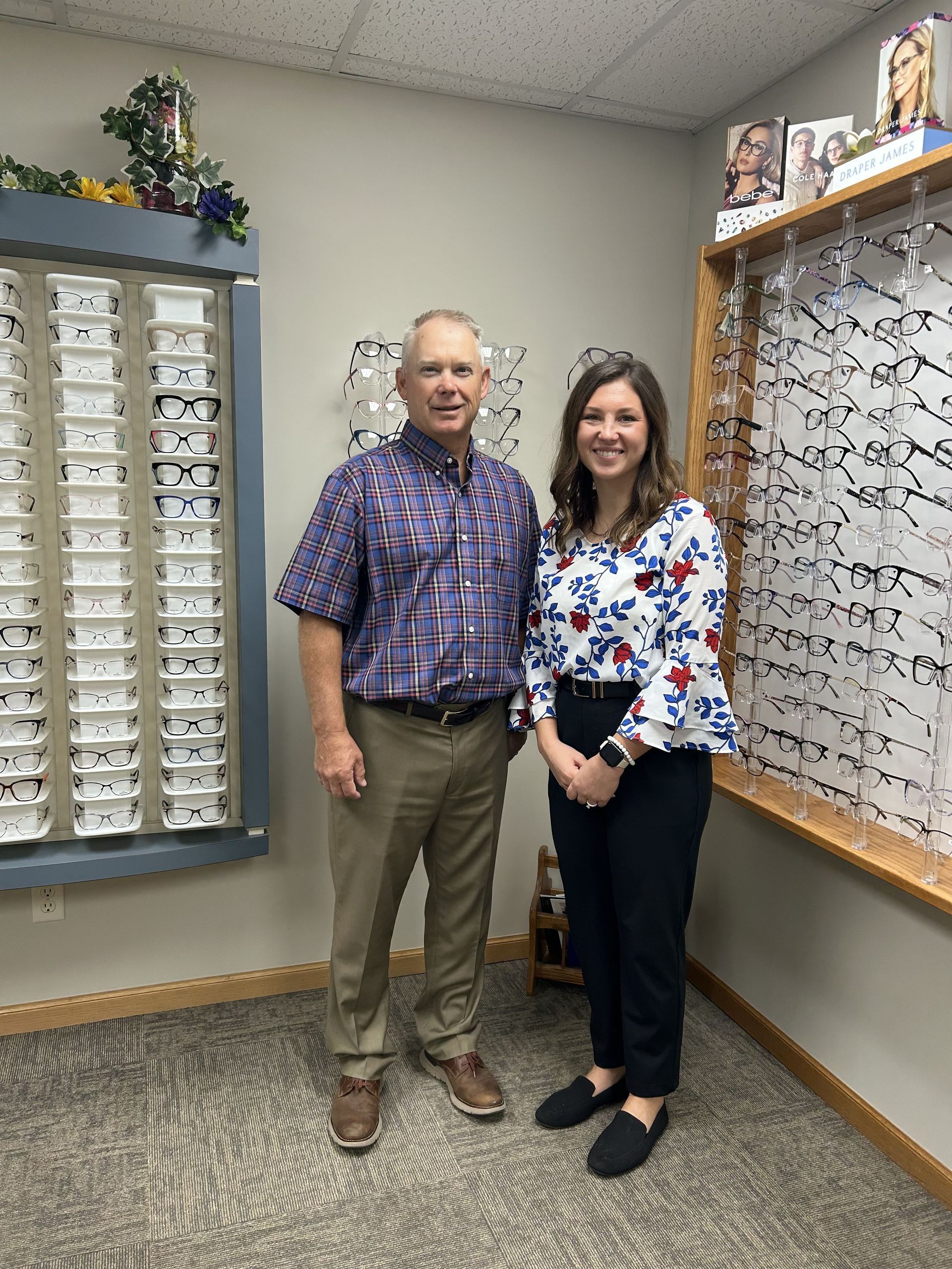 Two people standing in an eyeglasses display room, smiling beside rows of frames.