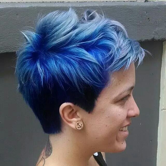 A woman with short blue hair is smiling and wearing earrings.