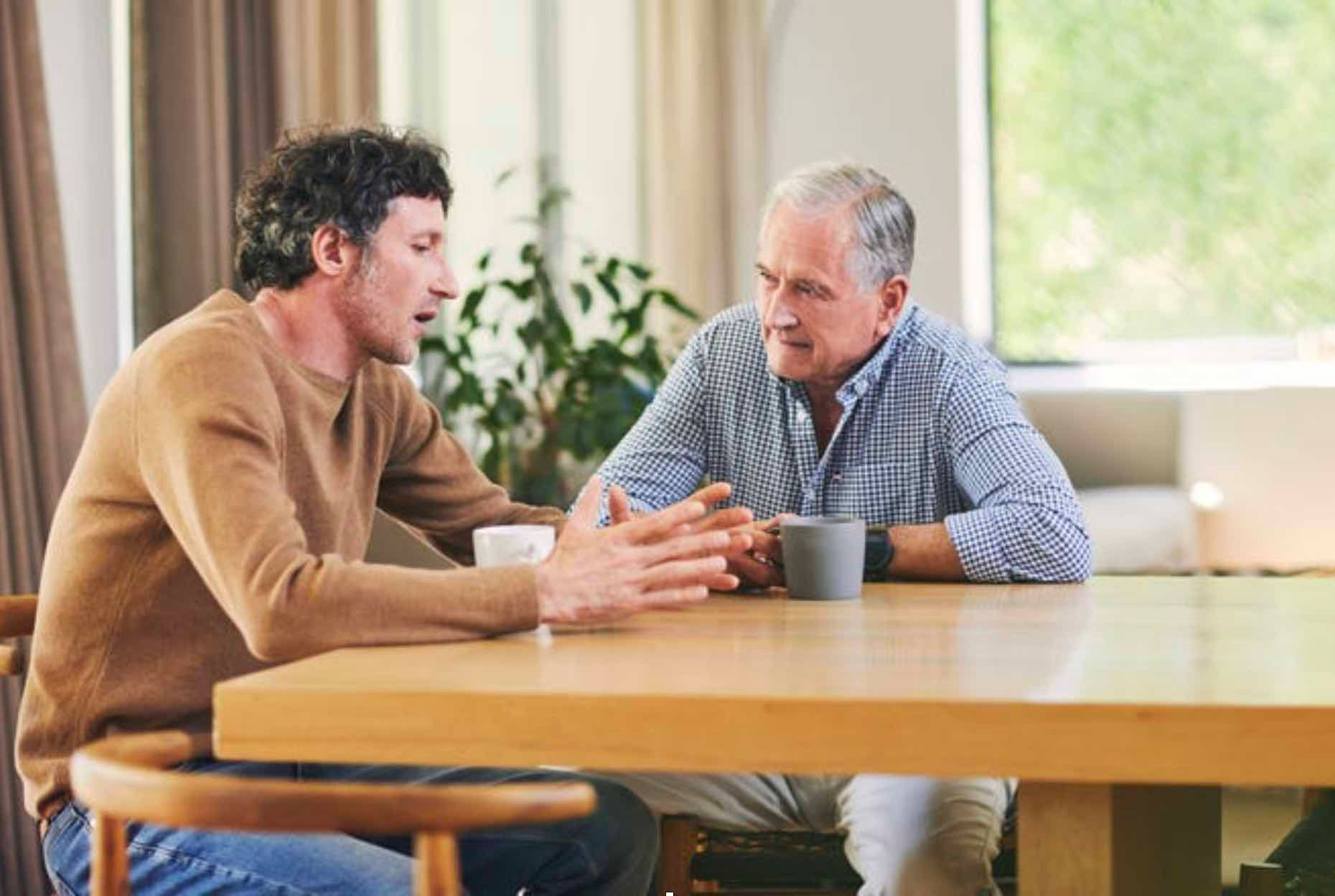 Two people talking at a wooden table in a bright café, one gesturing while the other listens.