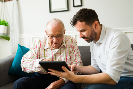 Two men sit on a couch, looking at a tablet together in a bright living room.