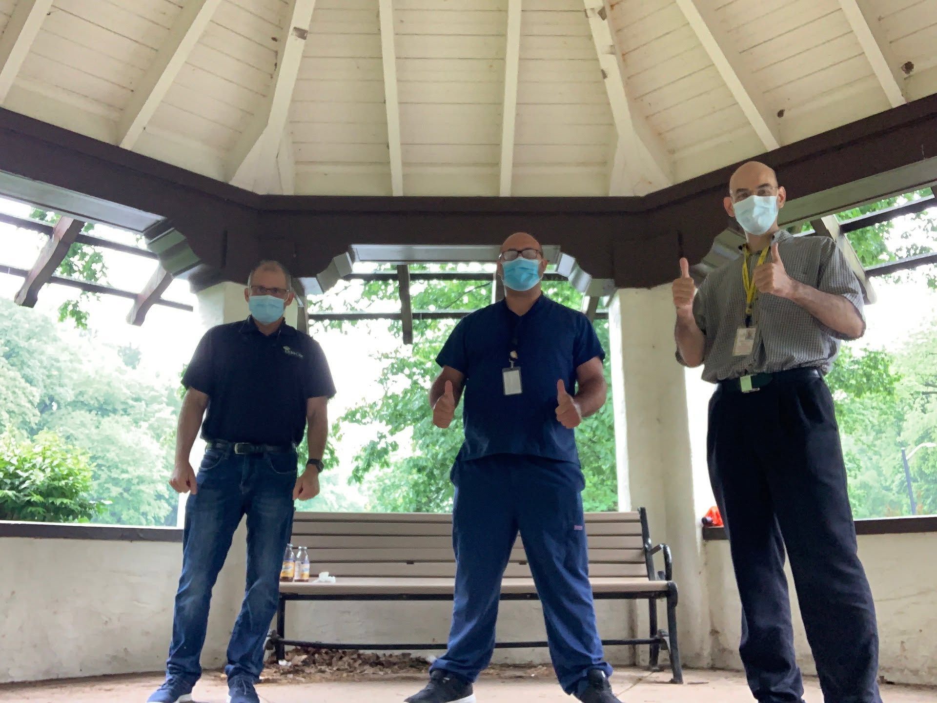 Three masked people standing in a gazebo, with benches and trees in the background.
