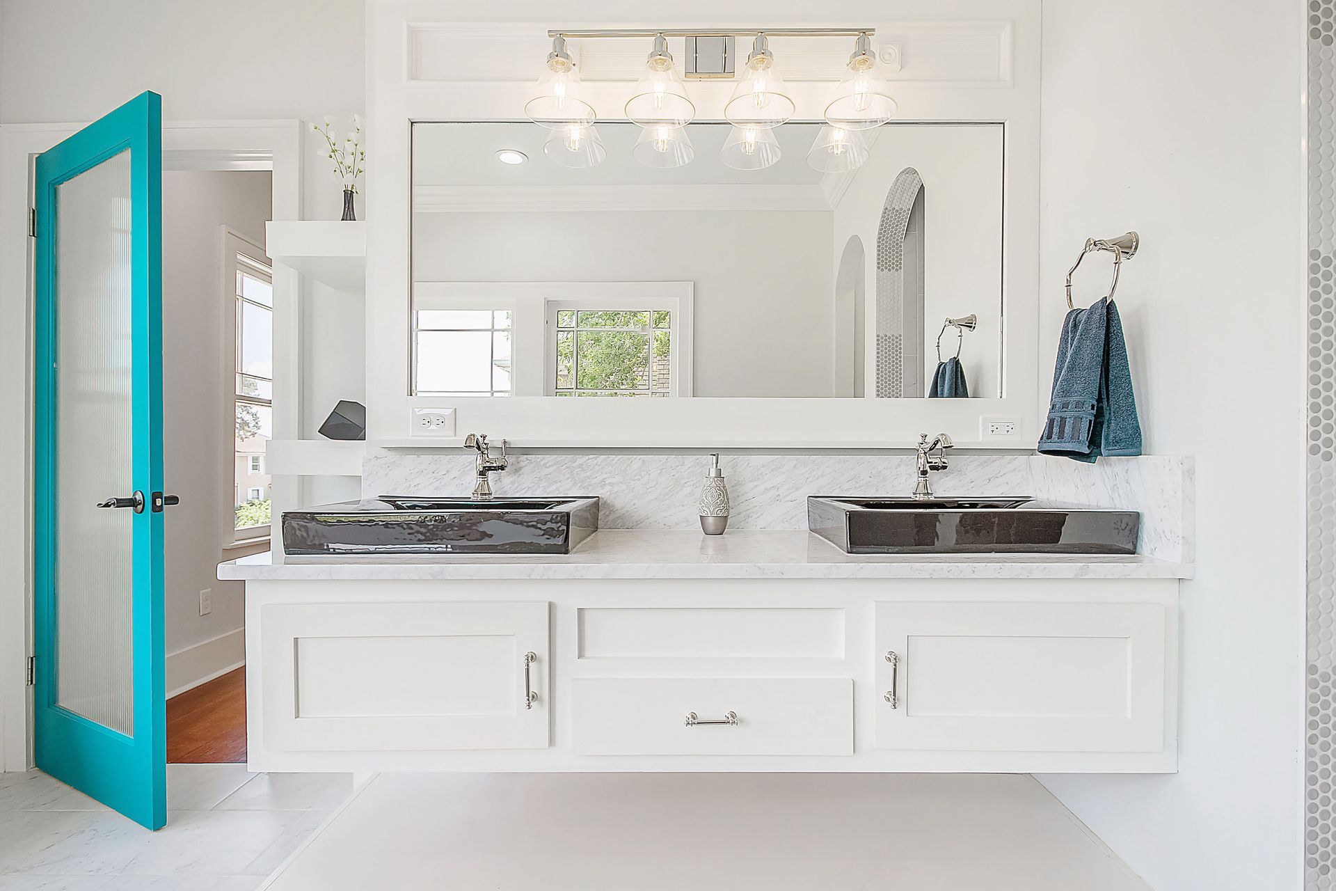 A bright, white bathroom with a turquoise door, a double vanity with black sinks, and a large mirror.
