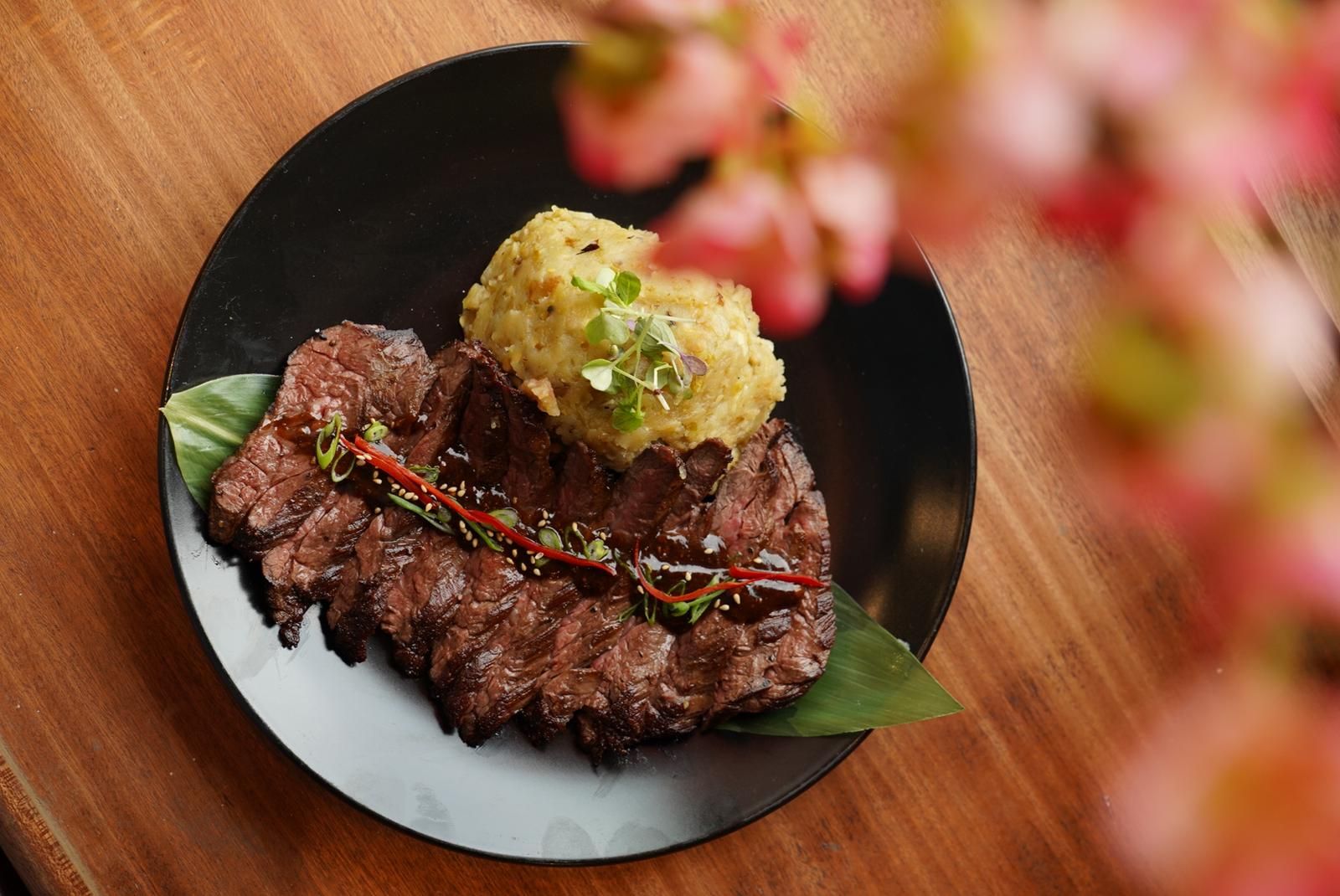Grilled steak topped with a mashed potato and sauce, served on a black plate.