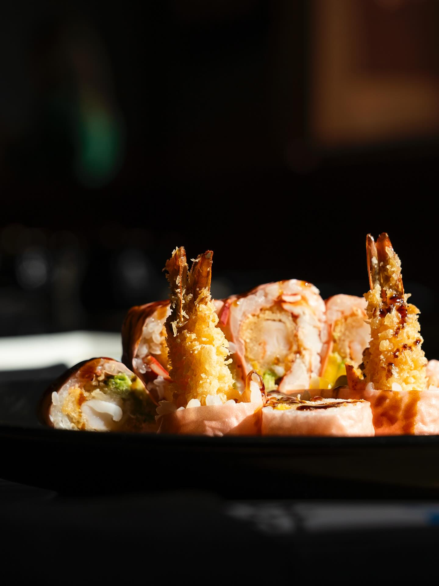 Sushi roll with fried shrimp on a dark plate, lit by sunlight.