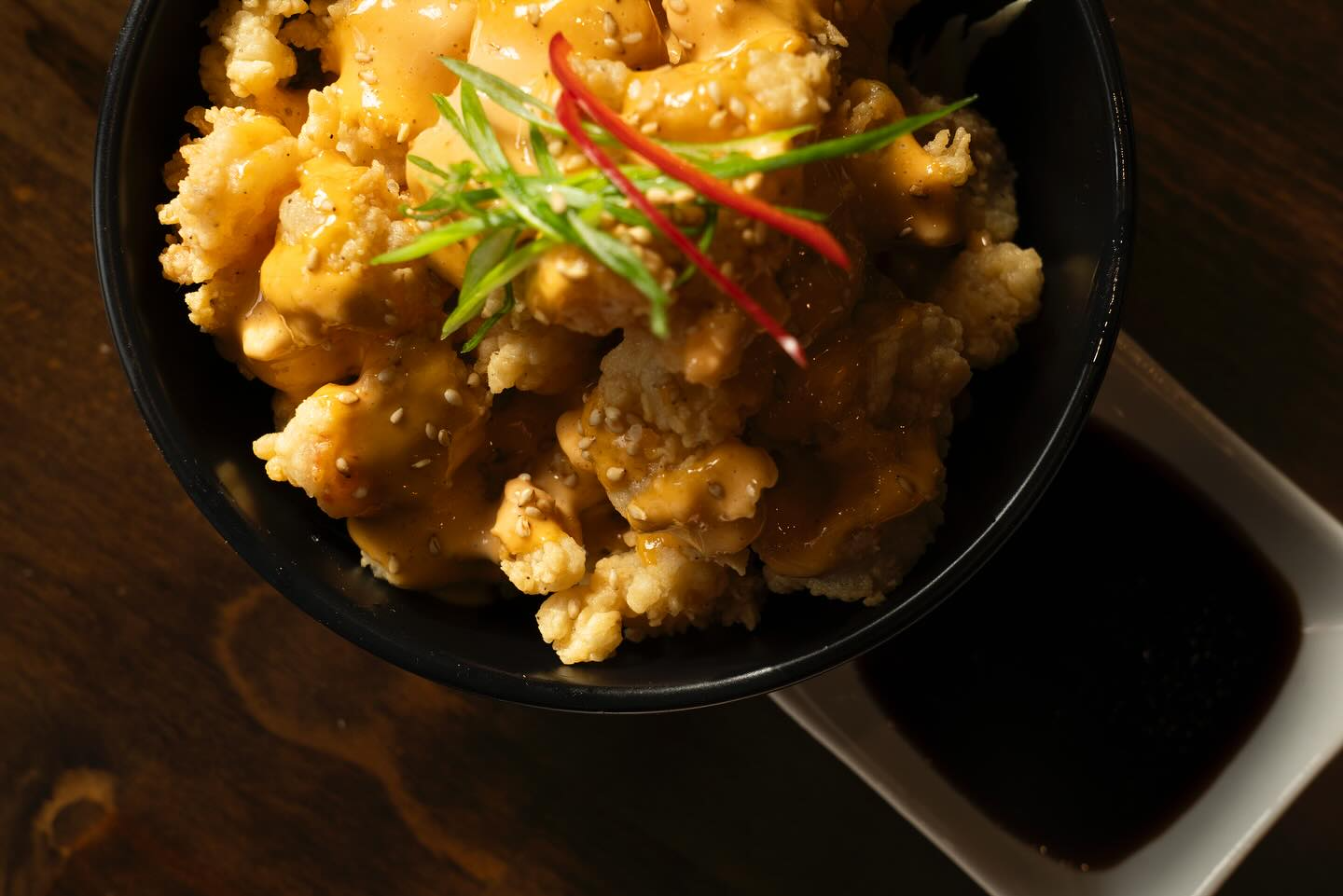 Fried food with orange sauce and scallions, served in a black bowl, with a side of sauce.