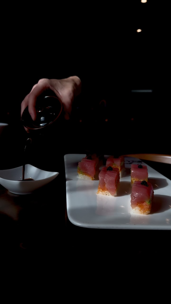 A hand pouring sauce over sushi bites on a white plate. Dark setting.