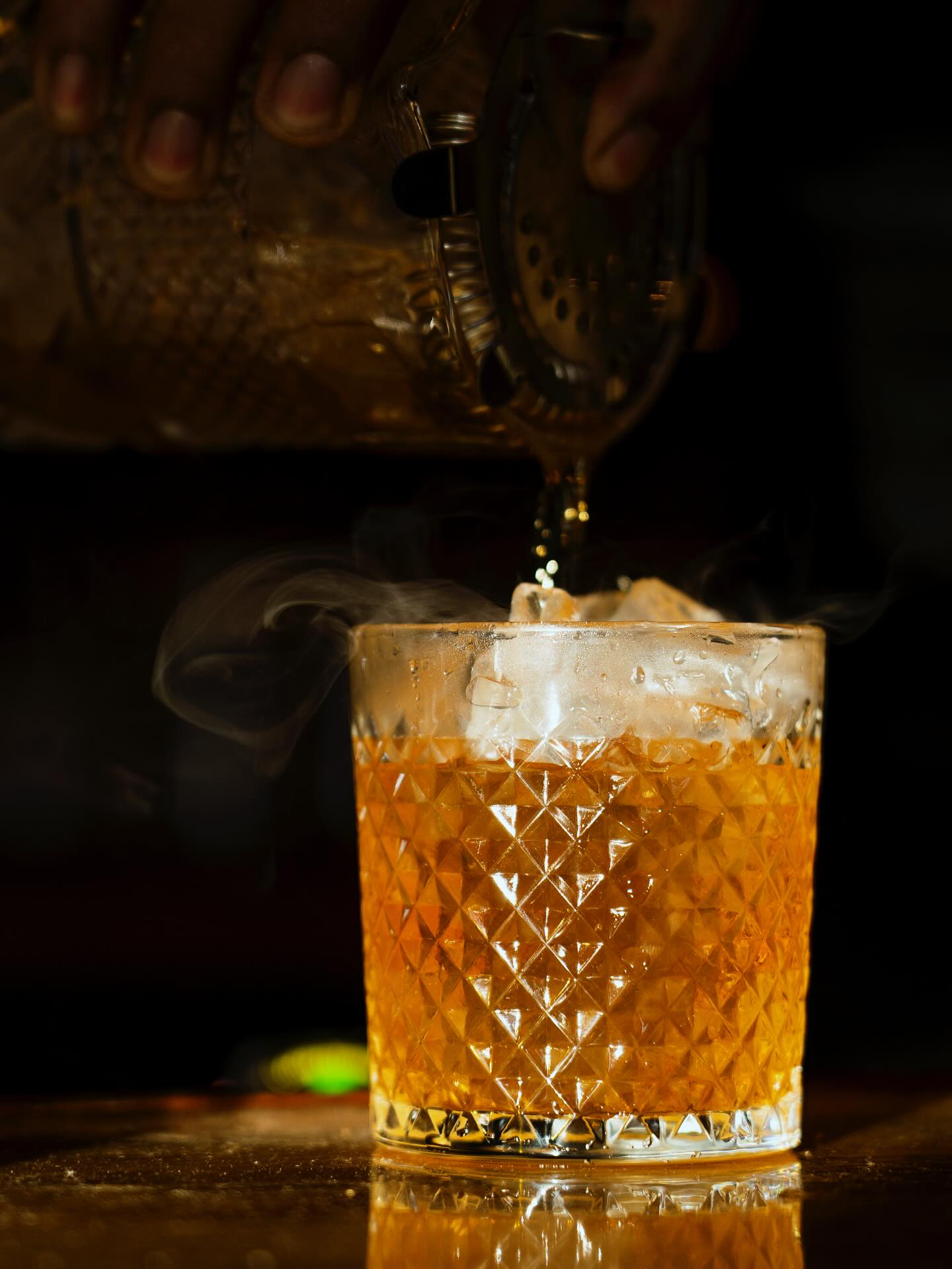 Cocktail being poured into a glass with ice; smoke rises. The glass sits on a wooden bar.