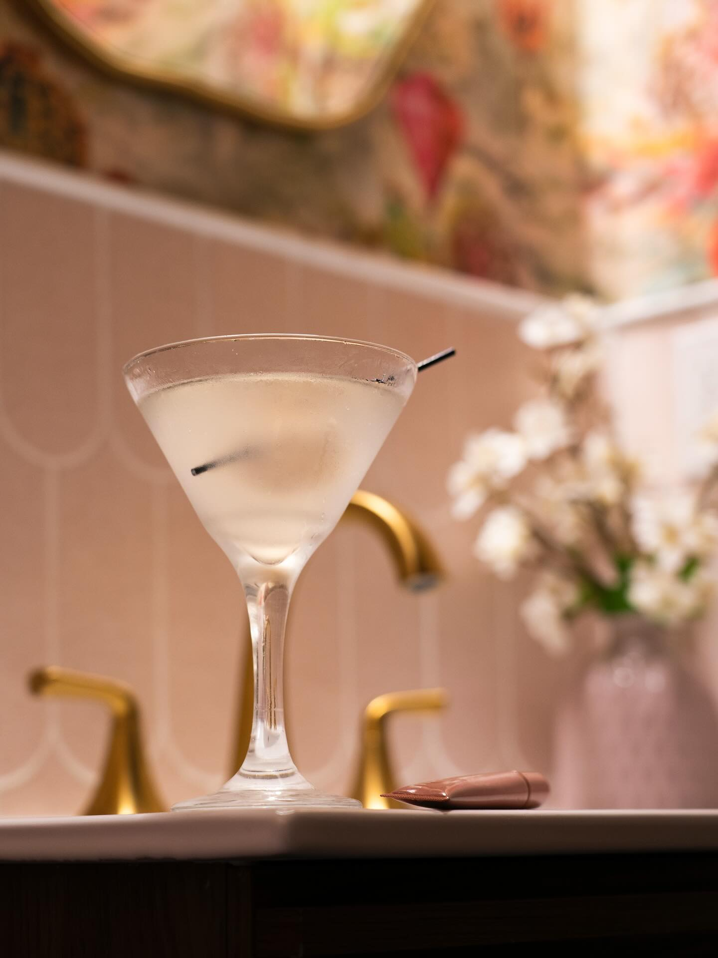 Martini in a glass on a bathroom sink, gold fixtures, flowers, and floral wallpaper in the background.