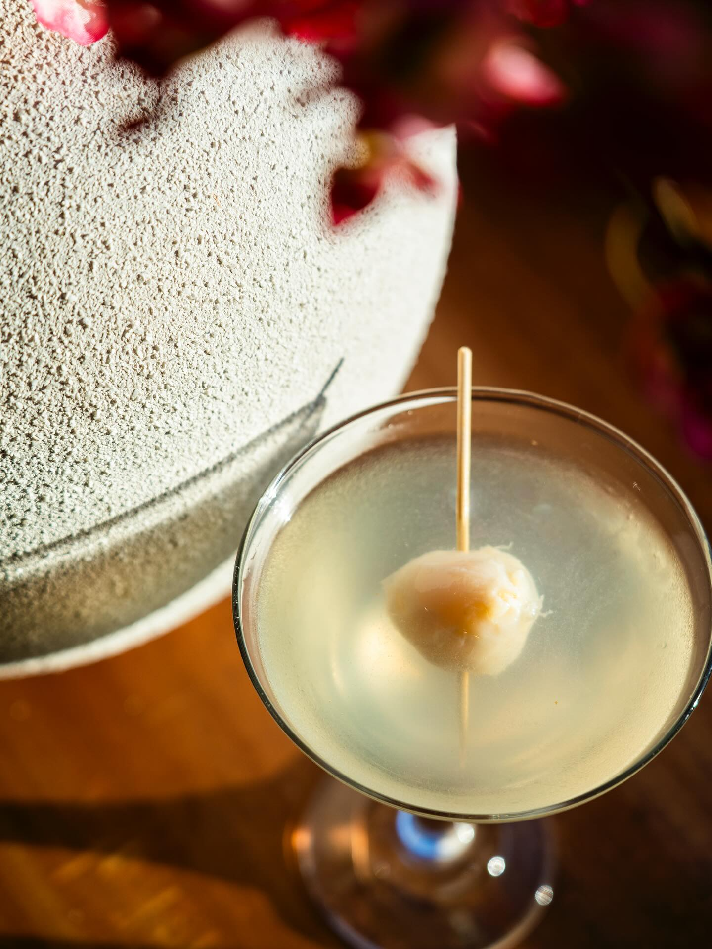 Cocktail in a coupe glass garnished with a lychee on a toothpick, near a decorative vase and flowers.