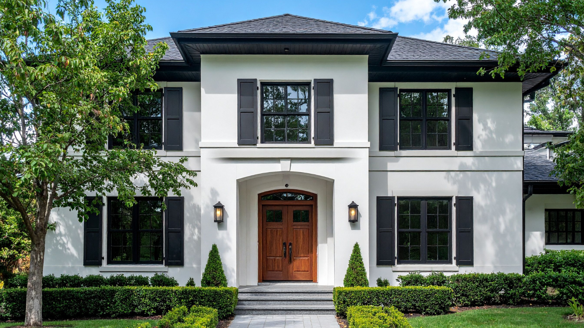 Two-story white house with black shutters, dark roof, and a wooden front door, framed by green landscaping.
