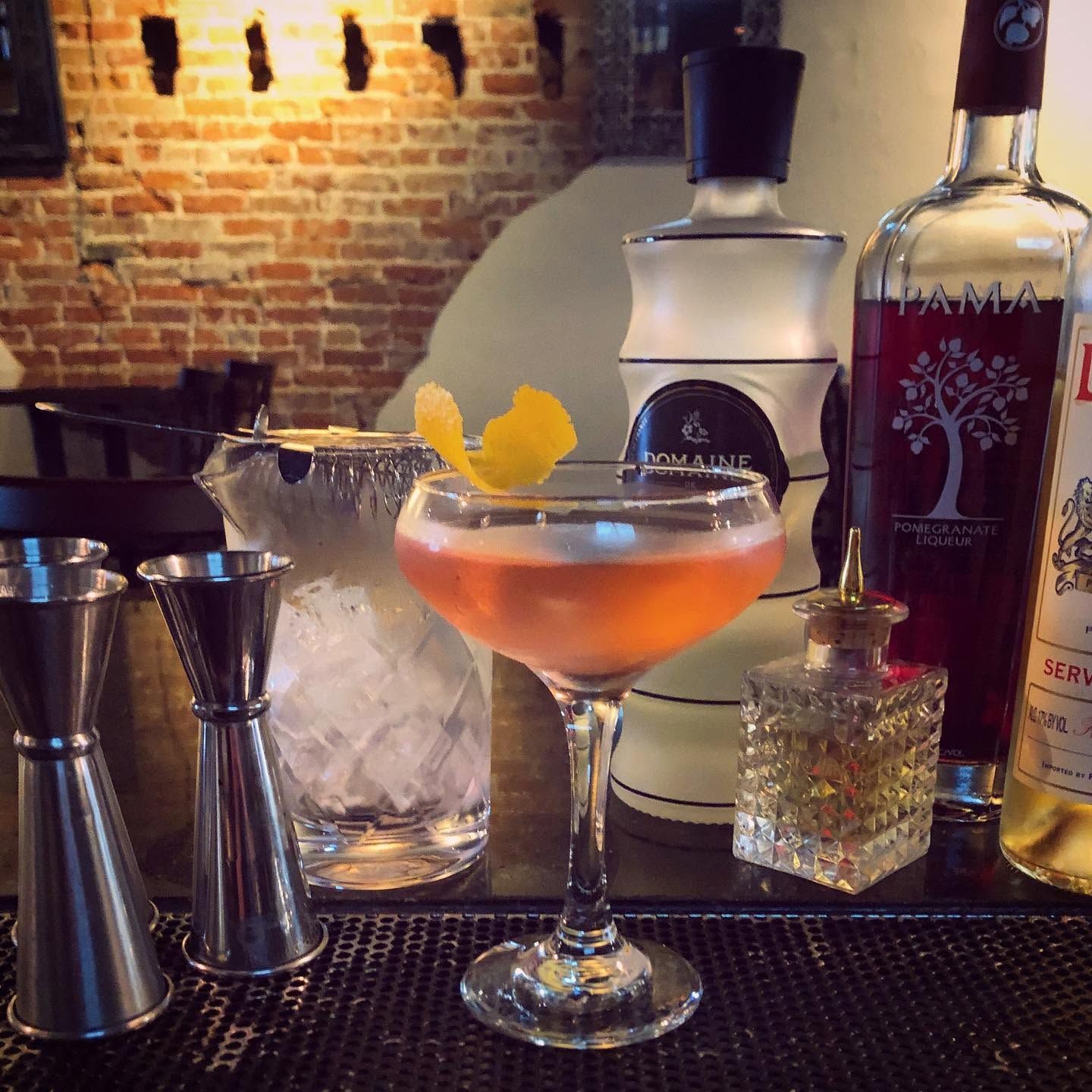 Cocktail in a coupe glass on a bar, with shakers, ice, and liquor bottles against a brick wall.