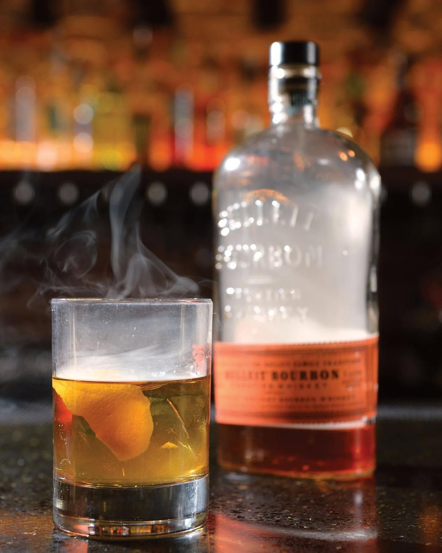 A whiskey glass with ice beside a bourbon bottle on a bar, with steam rising from the drink.