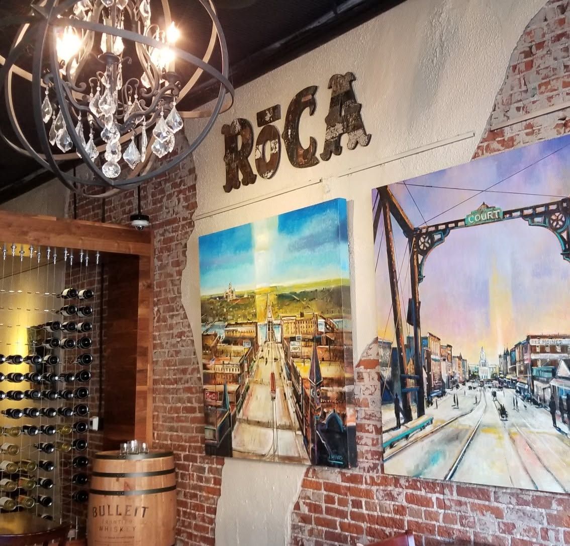 Rustic restaurant wall with “ROCA” sign, brick, hanging lights, and framed cityscape art.