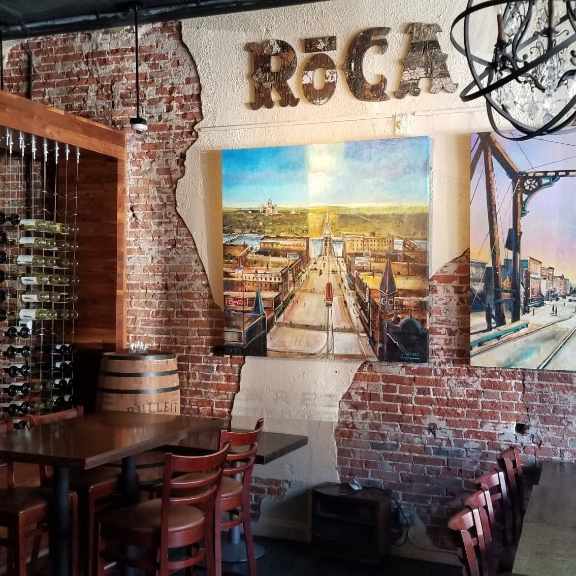Brick-walled restaurant interior with wooden tables and large wall art, including a cityscape and the word “Roca.”