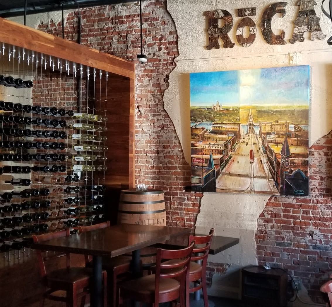 Wine bar interior with brick walls, bottle racks, wooden tables, and a cityscape painting.