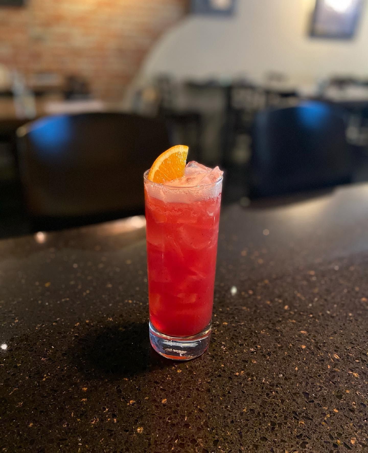 Tall red cocktail with an orange garnish on a dark bar counter, blurred lounge seating in the background