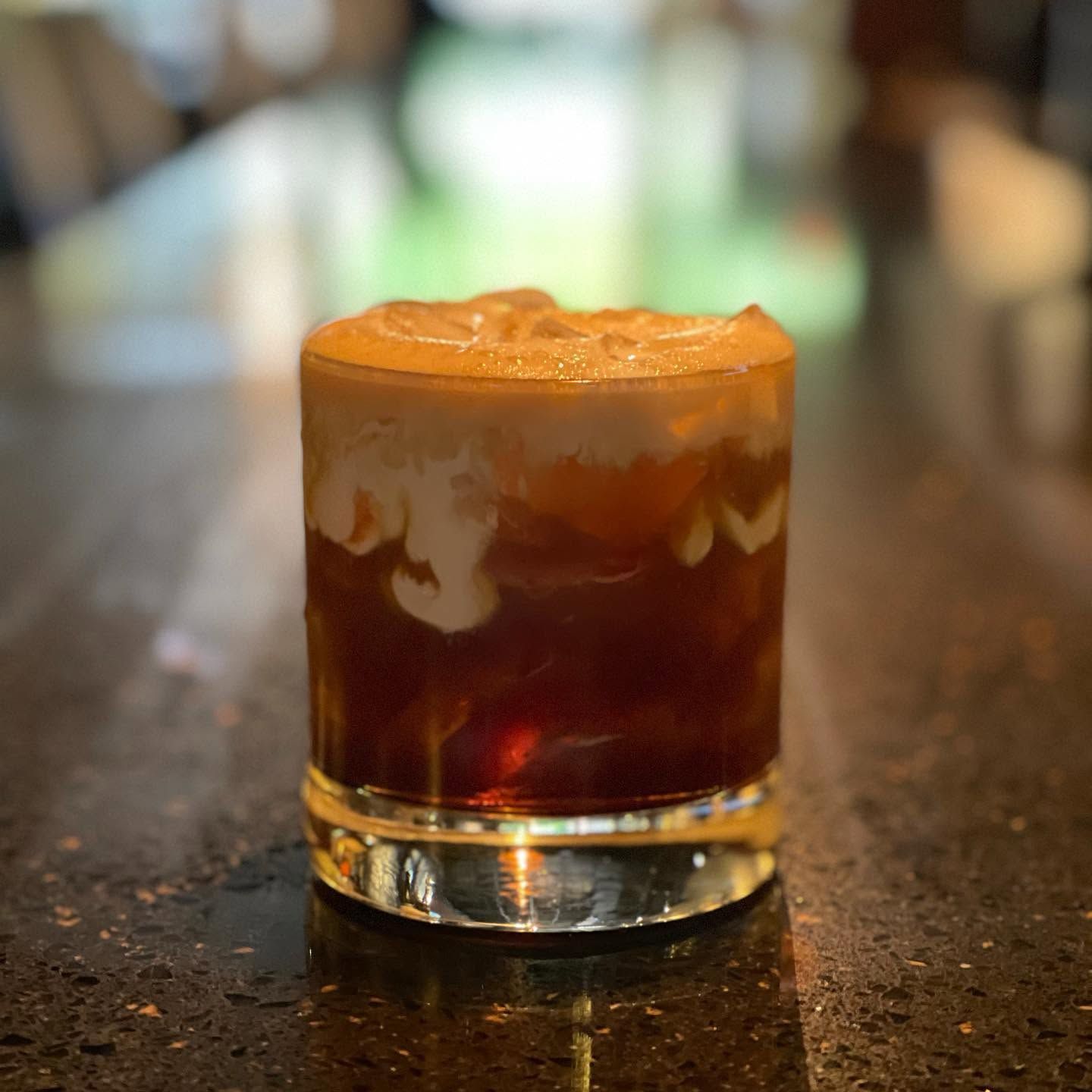 A small layered cocktail in a short glass on a bar counter, with a foamy top and blurred lights behind.