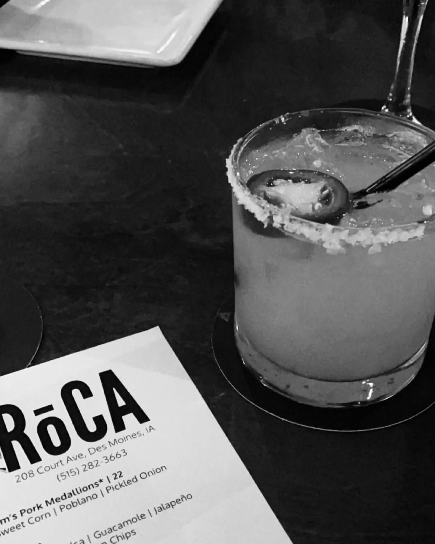 A salt-rimmed cocktail with a straw beside a RoCA menu on a dark table.