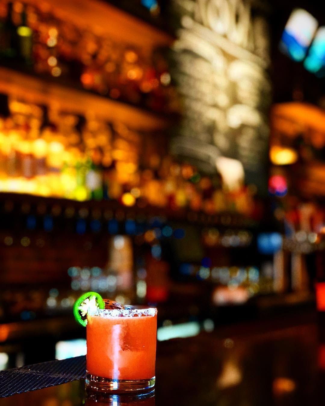 Orange cocktail with a lime garnish on a bar counter, with blurred liquor shelves in the background