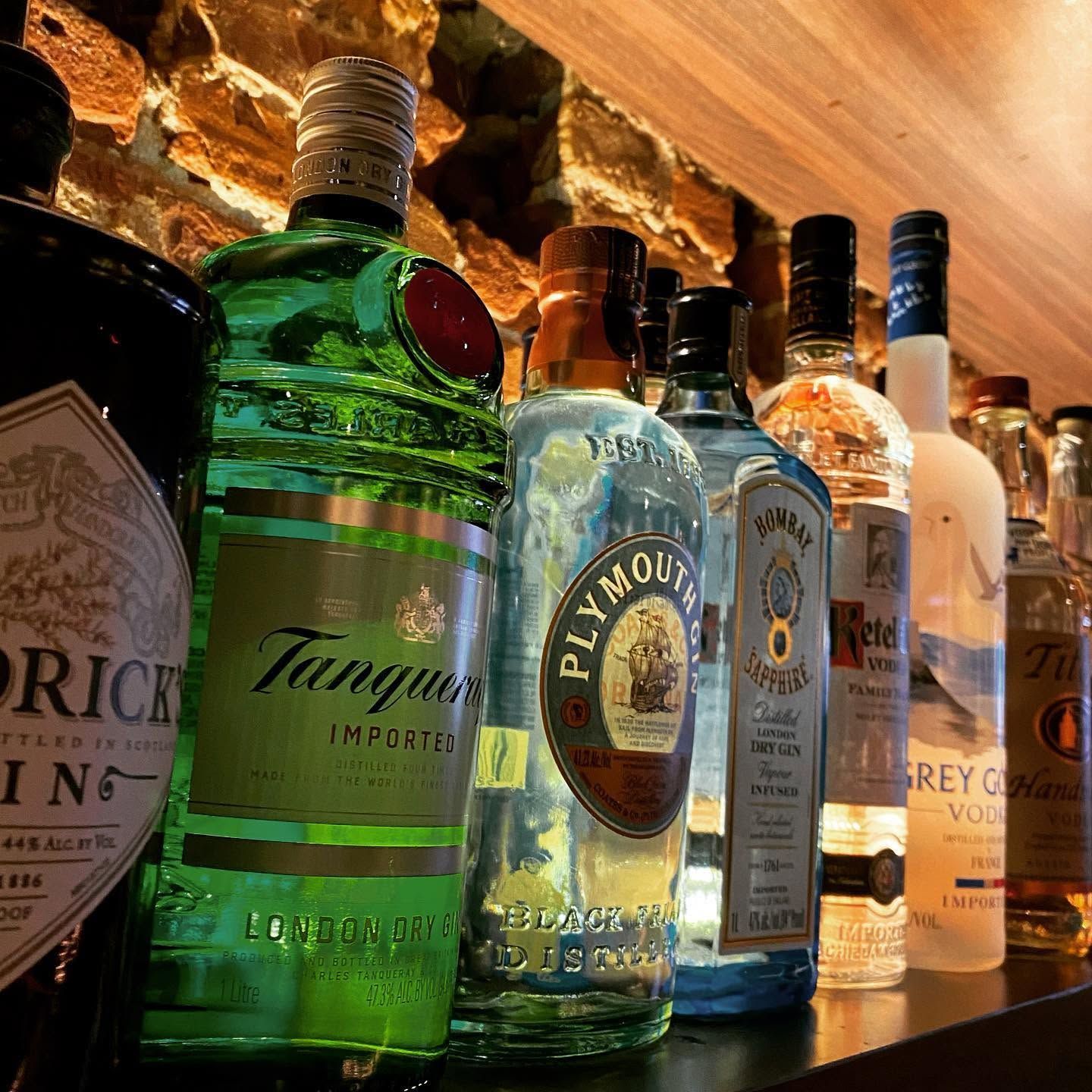 Row of gin bottles on a backlit bar shelf, including green Tanqueray and Plymouth bottles