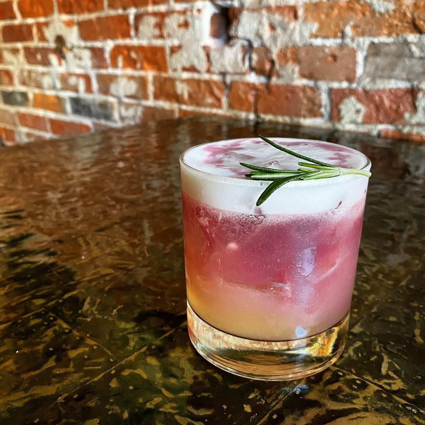 Pink and white layered cocktail with rosemary garnish on a textured wooden table against a brick wall.