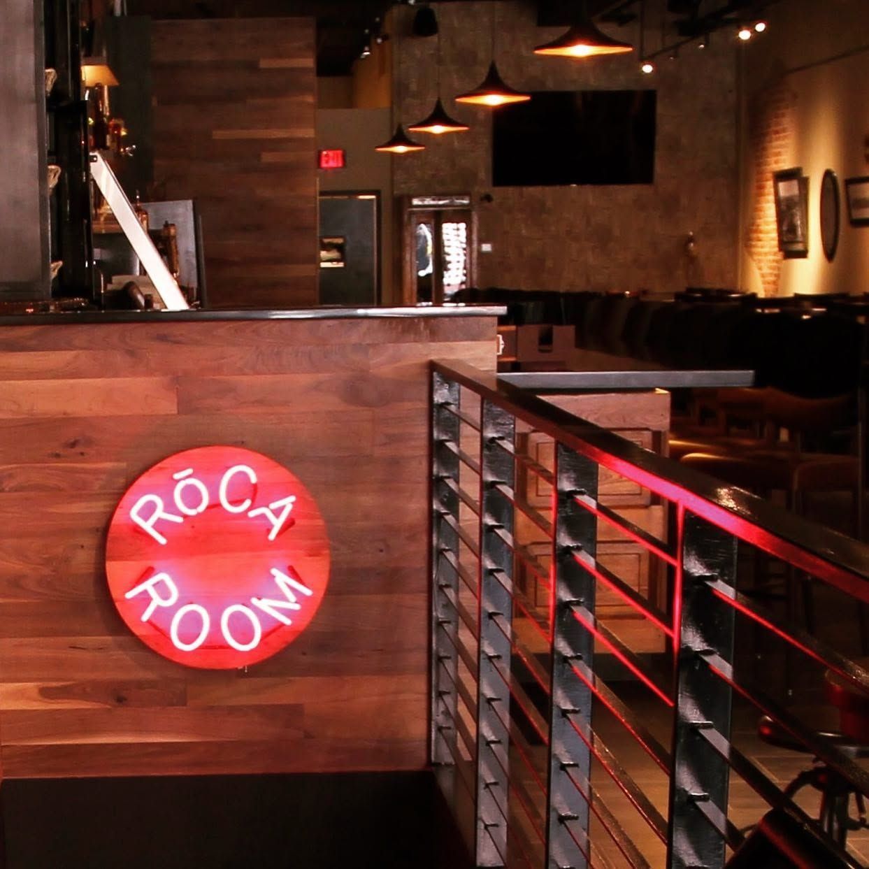 Dimly lit restaurant entrance with a red “Roca Room” sign and wood-paneled railings