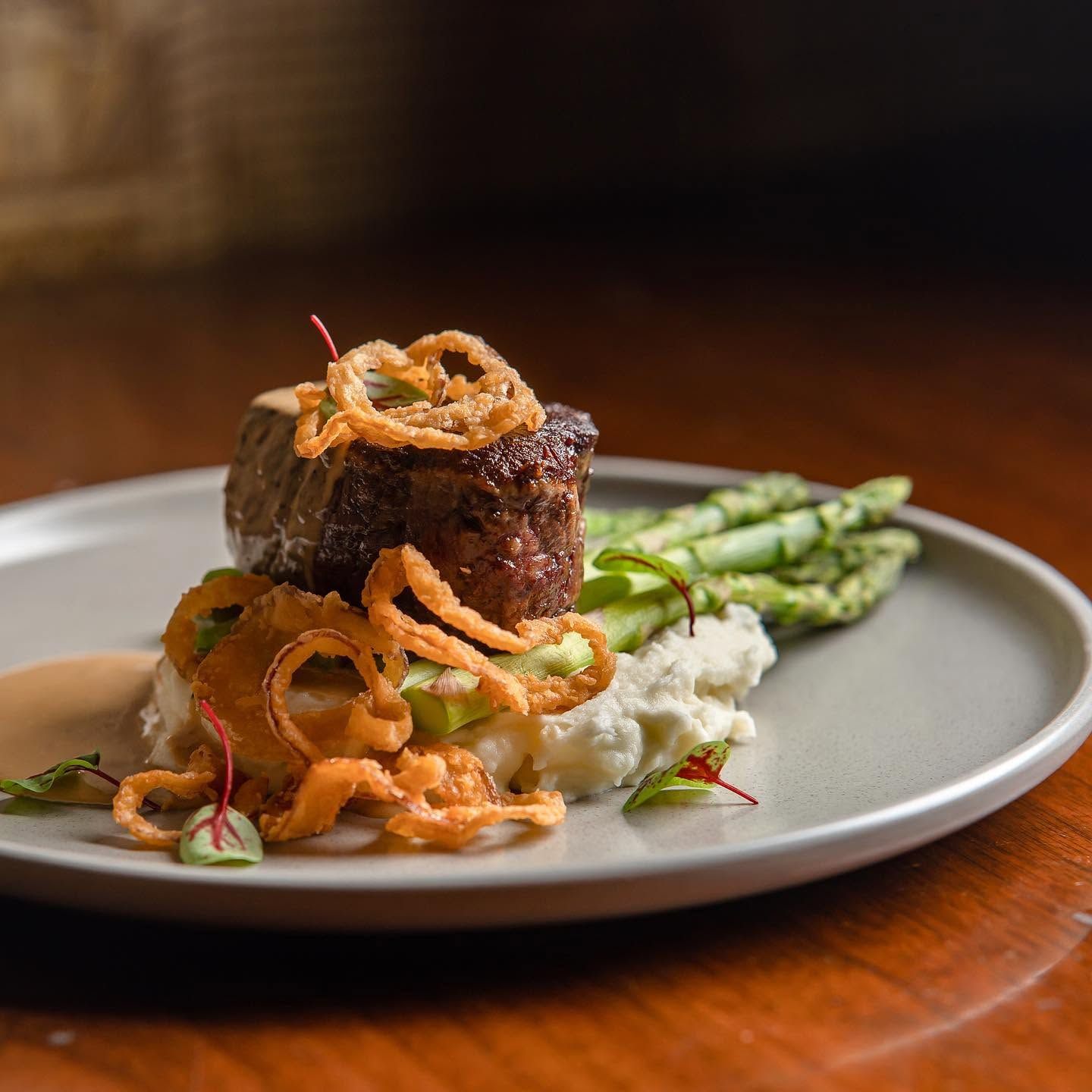 Plated gourmet steak with asparagus, mashed potatoes, and fried onion garnish on a white dish