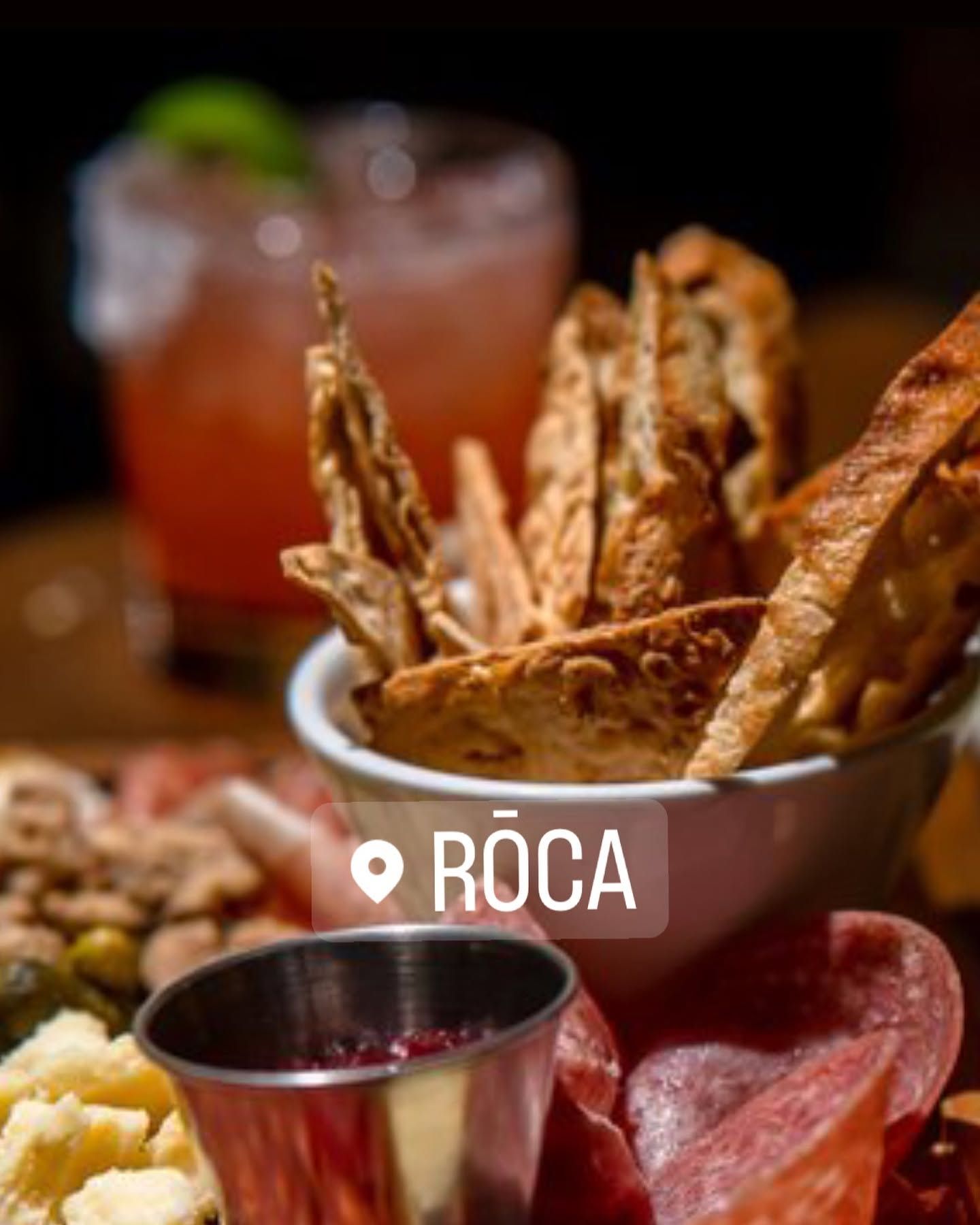 Breadsticks in a white bowl with dipping sauce and a drink, labeled “RÒCA” on a dark table.
