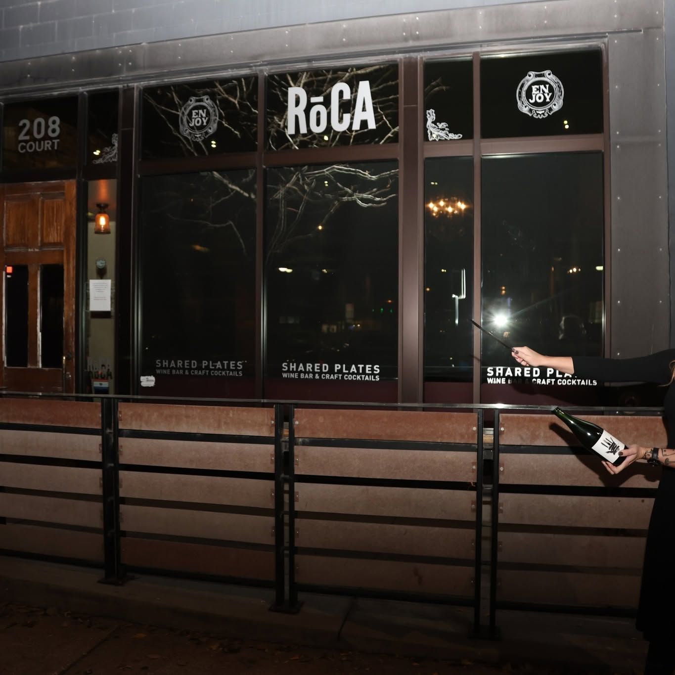 Nighttime storefront for RoCA with dark windows, lit signage, and a person holding a phone at the right edge