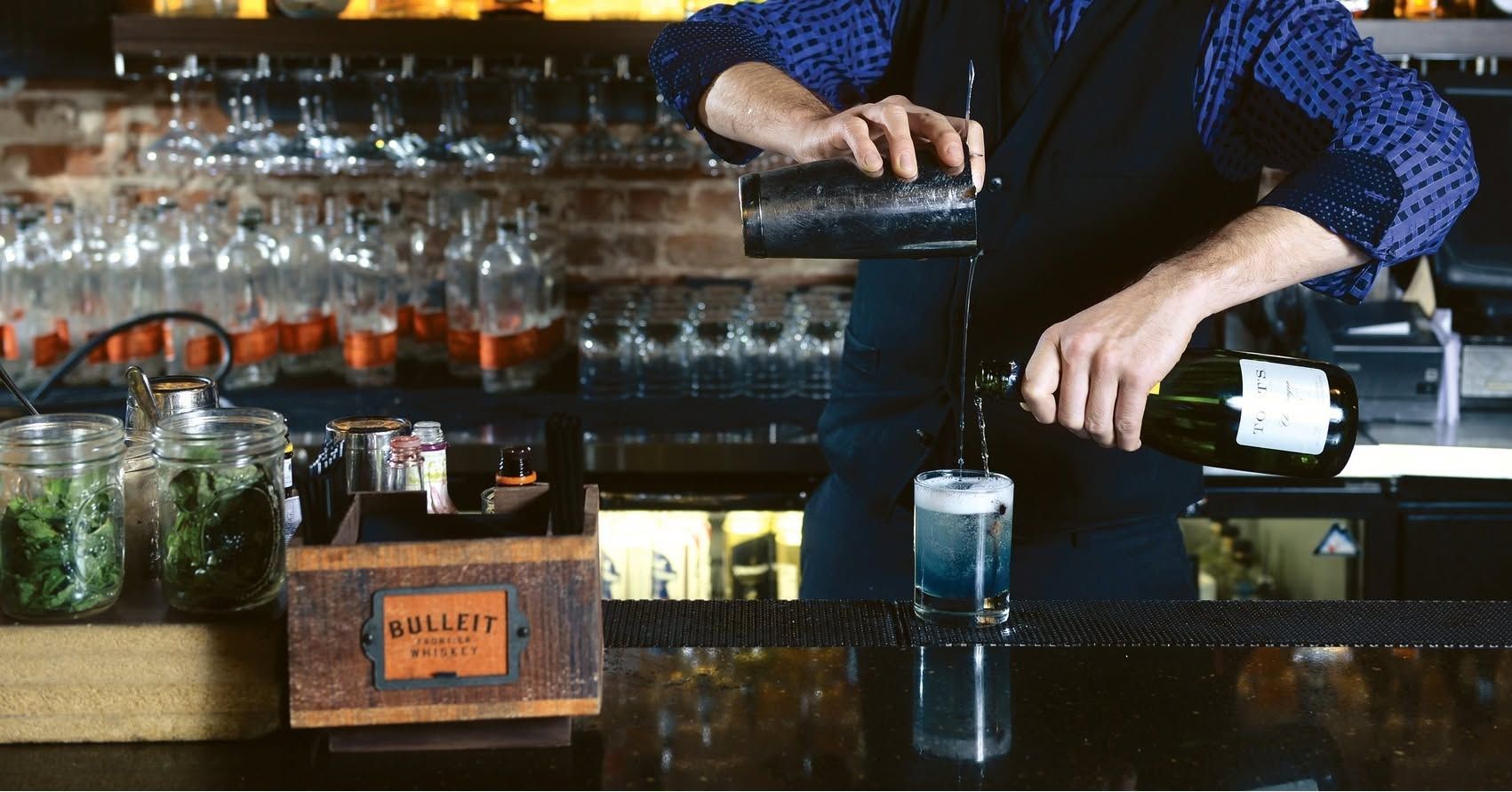 Bartender pouring a drink in a dim bar, with bottles and glassware behind the counter.