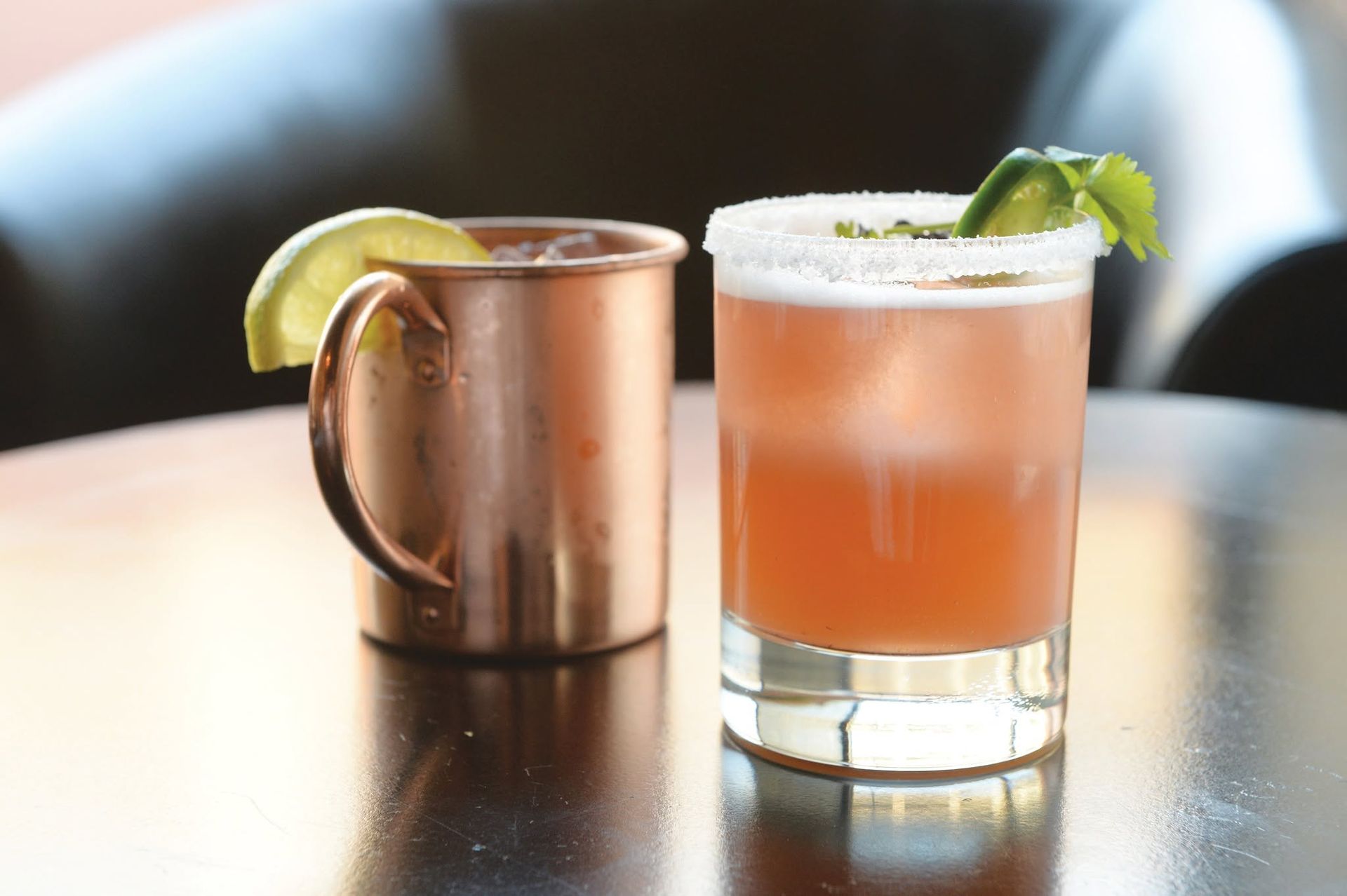 Two cocktails on a table: a copper mug with lime and a pink drink with a salt rim and mint garnish