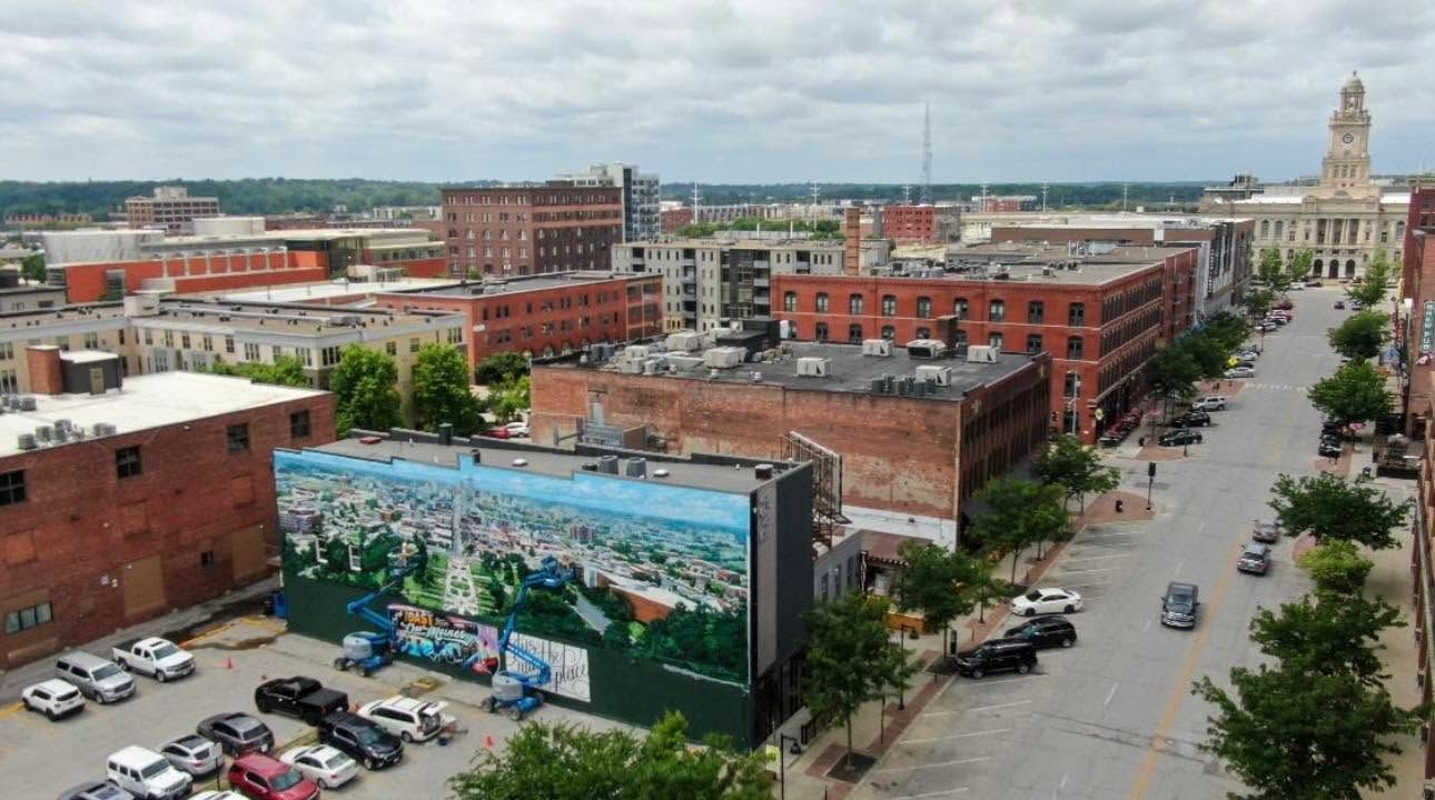 Urban aerial view of brick buildings, street, parked cars, and a colorful mural under a cloudy sky