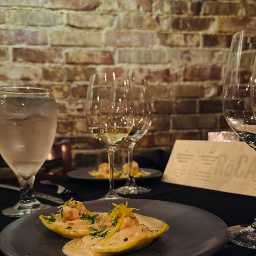 Restaurant table with wine glasses and a black plate of plated appetizers against a brick wall.