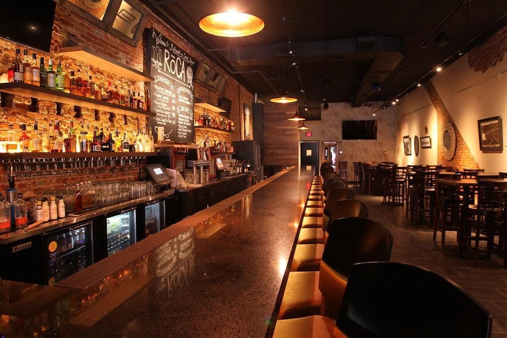 Dimly lit bar interior with long counter, rows of liquor bottles, and empty tables and stools