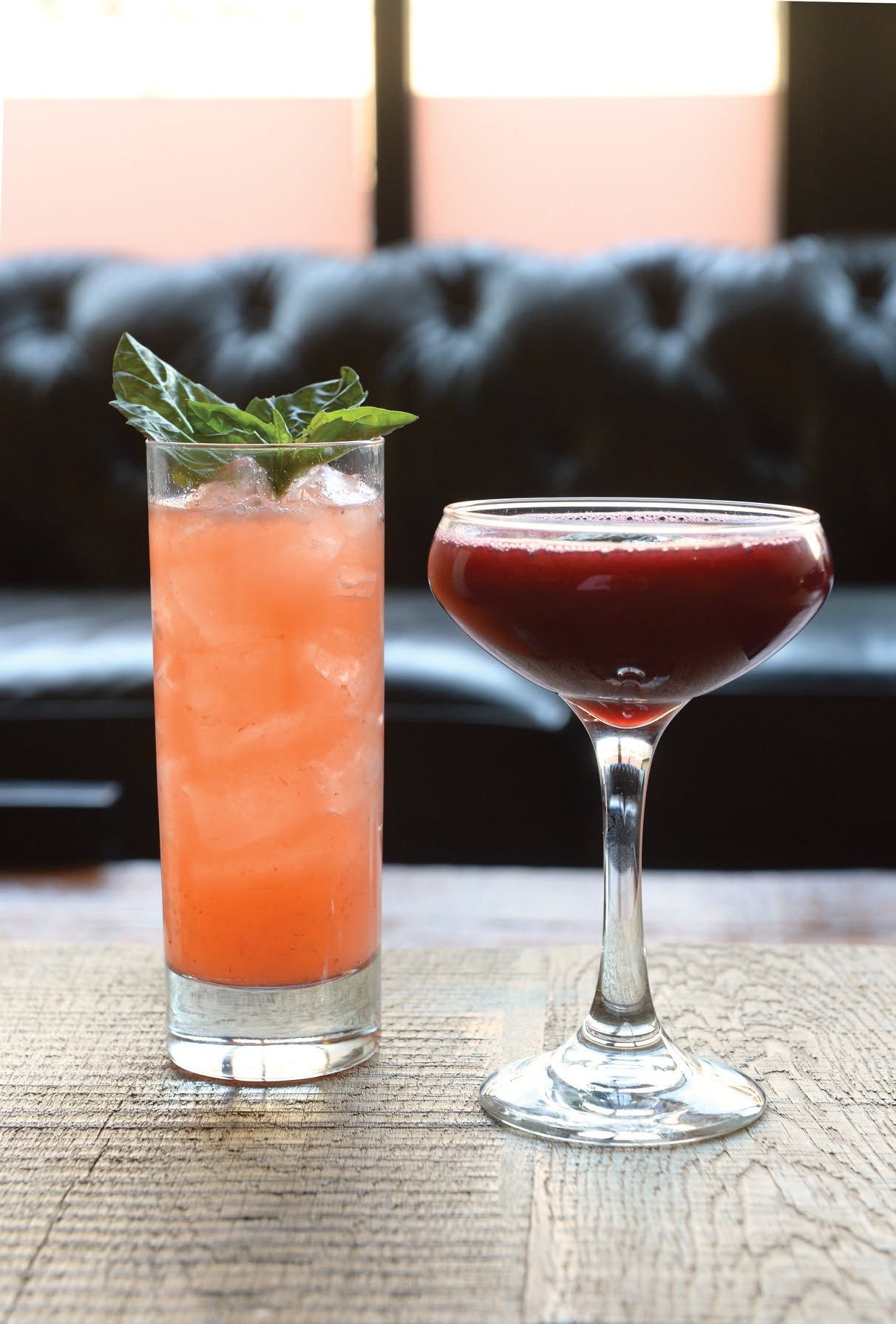 Two cocktails on a table: a tall orange drink with mint and a dark red martini in a stemmed glass.