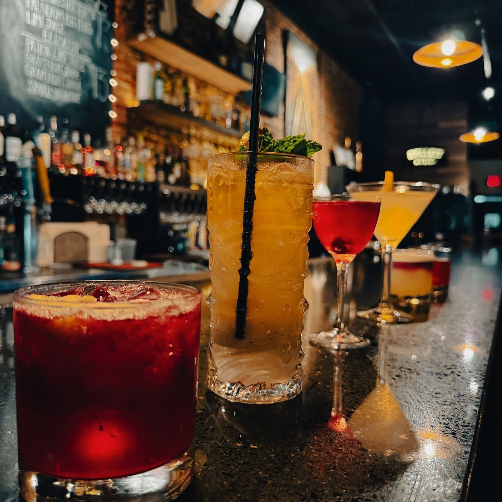 Colorful cocktails lined up on a dimly lit bar counter in a cozy nightlife setting