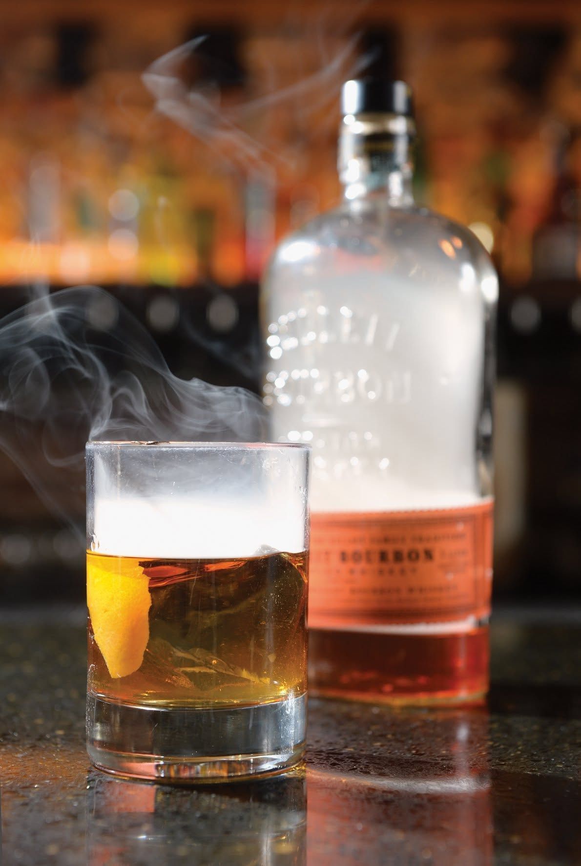 Cocktail glass with orange peel and rising vapor beside a frosted liquor bottle on a bar